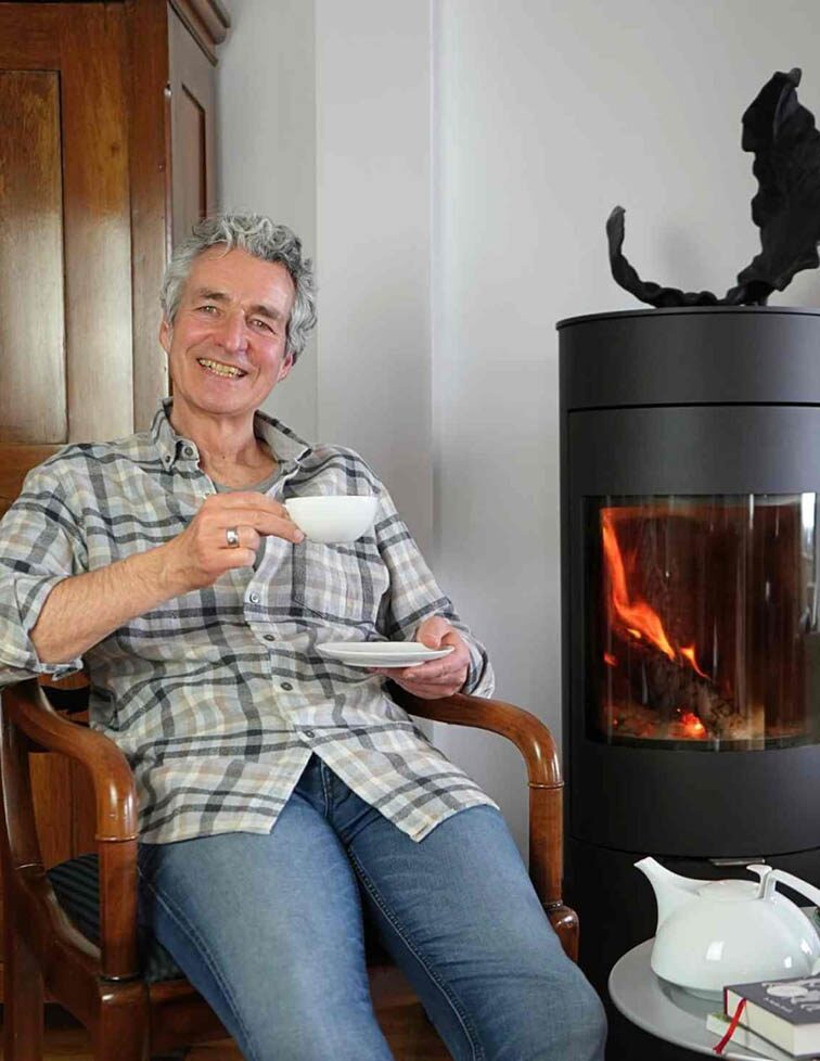 Mann sitzt lächelnd mit Teetasse im Sessel vor einem brennenden Kaminofen, daneben eine Teekanne