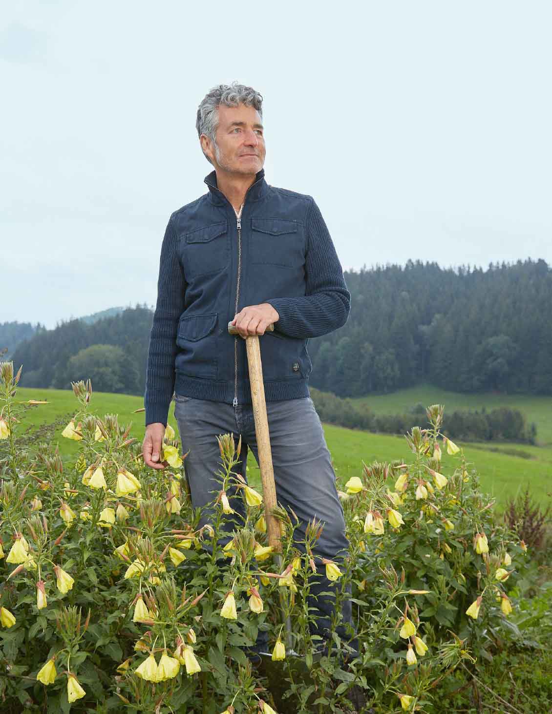 Mann in dunkler Jacke stützt sich auf einen Spaten, umgeben von hohen Stauden mit gelben Blüten