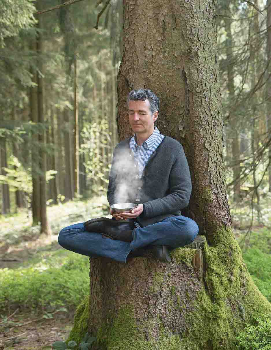 Ein Mann sitzt im Schneidersitz an einem Baumstamm im Wald, mit dampfender Schale in den Händen