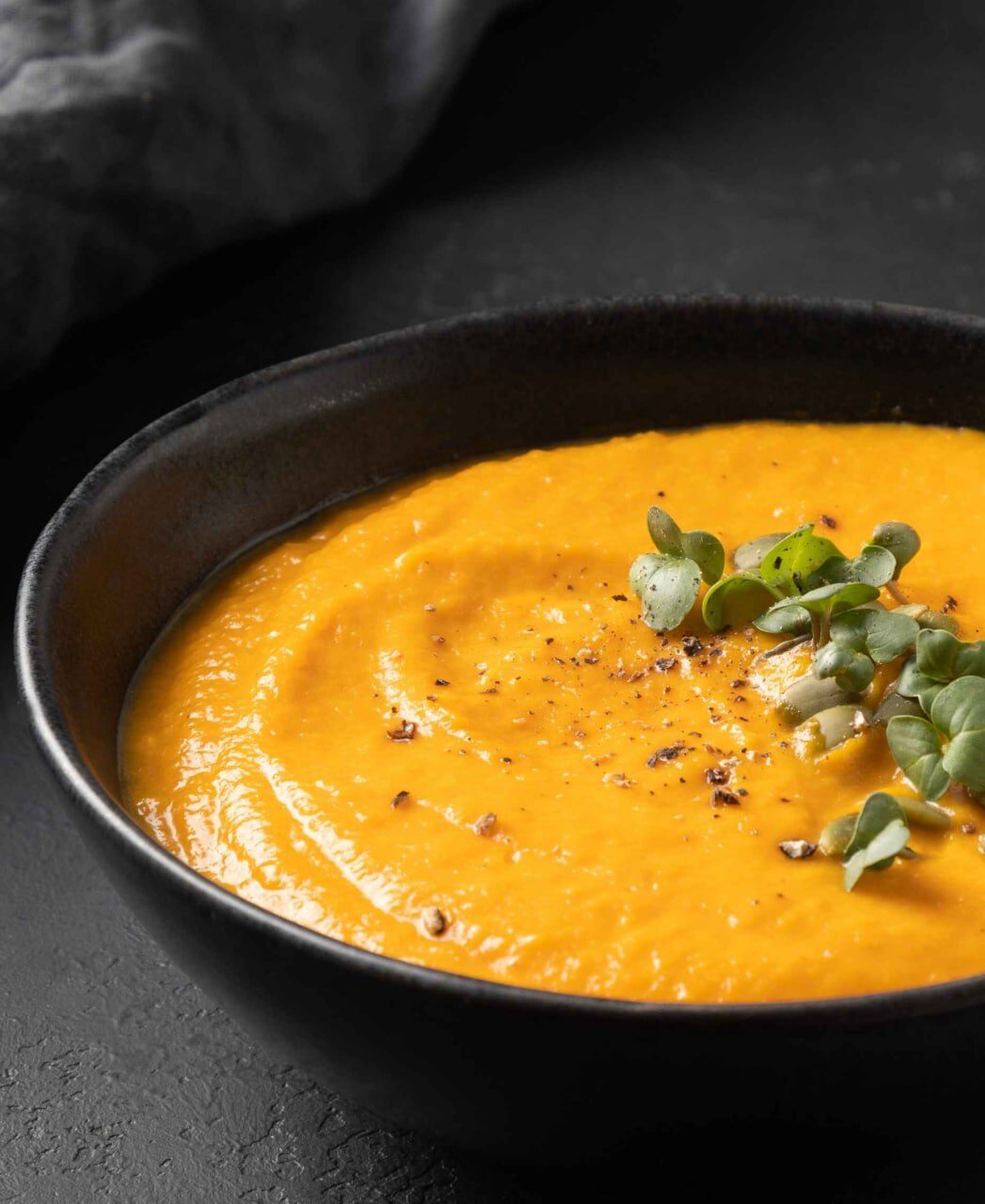 Orange Cremesuppe in dunkler Schale, mit frischen Keimlingen und grobem Pfeffer bestreut